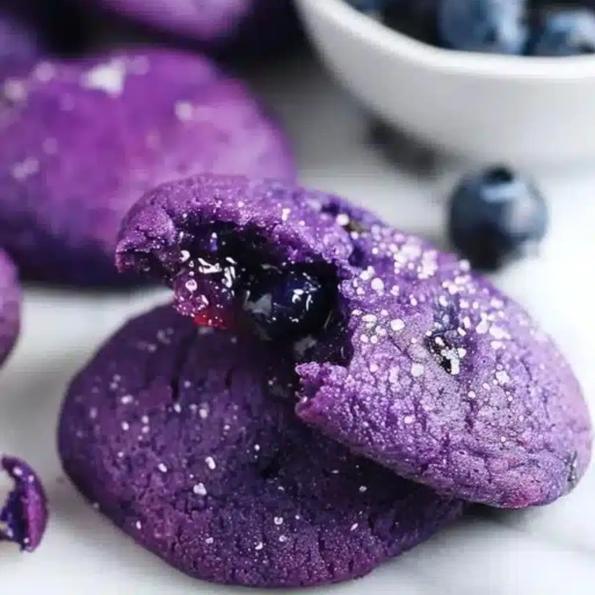 Chocolate cookies with blueberry jam centers arranged on a white plate, showing one cookie broken in half to reveal the purple jam filling inside, with sugar crystals visible on the cookie surface