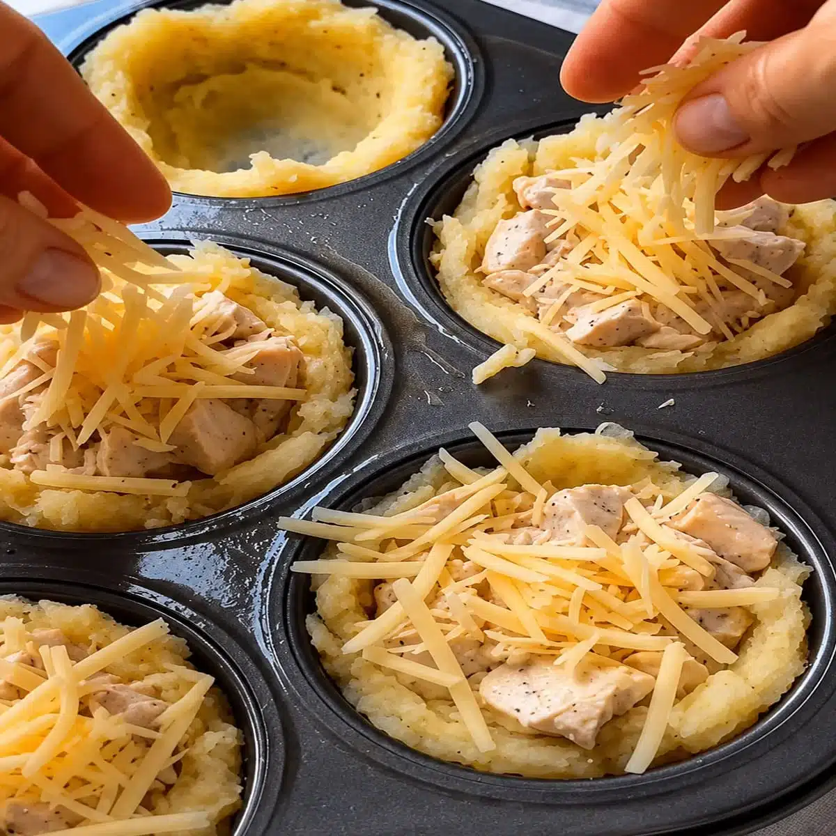 Cheesy Chicken Potato Nests – Easy Chicken Appetizer 6 Cheesy Chicken Potato Nests being filled in a muffin tin with creamy chicken pieces, shredded cheese, and shaped potato cups before baking.